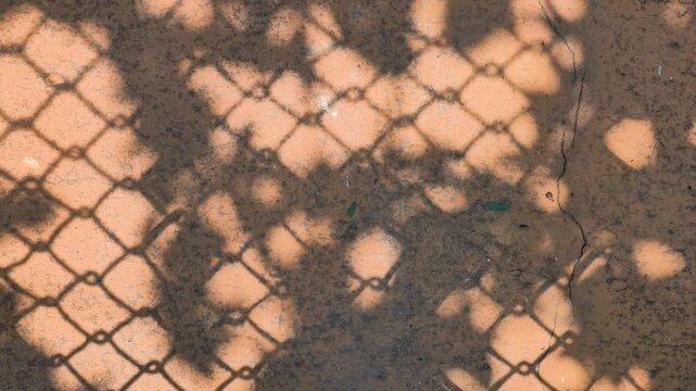 Top down view of chain link fence shadow moving over textured orange painted surface in bright sunlight.