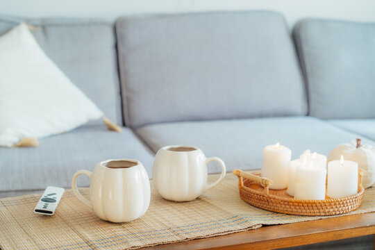 Autumn fall mood composition for hygge home decor. White pumpkin's shaped cups with hot drink, burning candles, remote controller on coffee table in living room. Movie night at home. Cozy relax time.