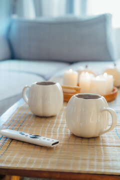 Autumn fall mood composition for hygge home decor. White pumpkin's shaped cups with hot drink, burning candles, remote controller on coffee table in living room. Movie night at home. Cozy relax time