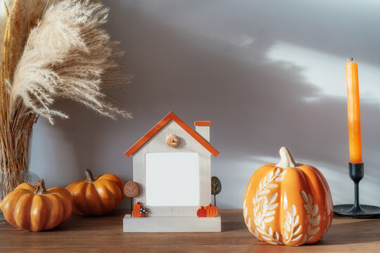 Cozy warm autumn home interior - decorative orange ceramic pumpkins, house's shaped photo frame with copy space, candles, dried flowers on the wooden console. Seasonal hygge home fall decor