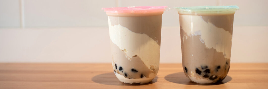 Two cups of bubble tea with tapioca pearls. One cup has a pink lid and the other a green lid. The drinks have a creamy texture and are placed on a wooden surface. banner, copy space
