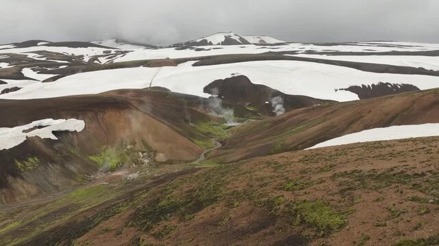 Drone pans left over the Laugavegur trail in Iceland, revealing a vast, snow-covered and rugged mountain landscape with visible steam vents and a winding stream. Hikers traverse the remote terrain.