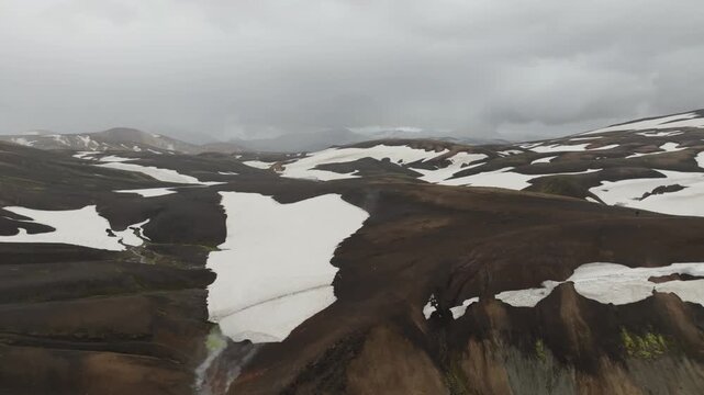 Aerial view of the unique landscape surrounding Stórihver spring on the Laugavegur hiking trail on Iceland. The drone pans right, revealing volcanic hills, snow patches, and steam vents.