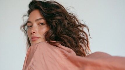 Fototapeta premium Portrait of a woman with flowing dark hair looking over her shoulder in soft pink clothing against a plain light background.