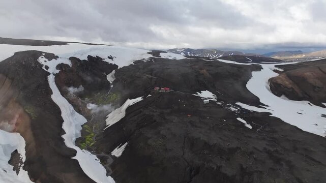 Aerial push in shot of the unique volcanic landscape on the Laugavegur hiking trail on Iceland. Discover hikers and the mountain hut Hrafntinnusker amidst snowfields and geothermal steam.