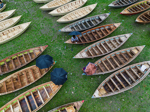 Aerial view of many wooden boats resting on a lush green field with people using umbrellas for shade in Manikganj, Dhaka Division, Bangladesh.