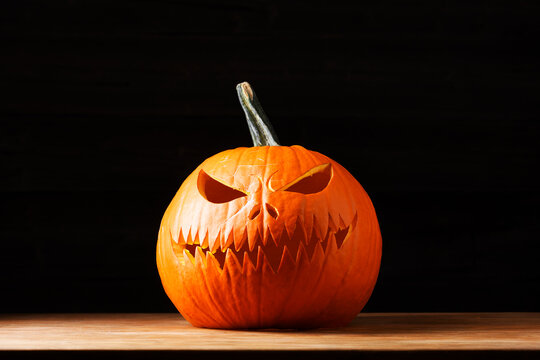 Carved Halloween pumpkin with an evil face placed on a wooden table against a black background. Traditional autumn decoration with copy space for seasonal holiday designs
