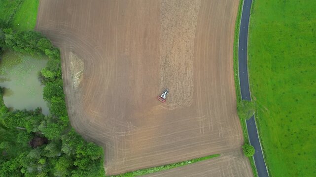 Top-down tractor plowing brown field next to pond and asphalt road