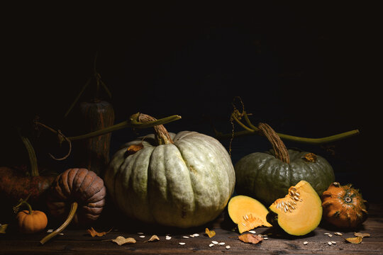 Assortment of pumpkins with cut pieces on wooden table in dark moody lighting. Autumn harvest composition with squash slices and rustic texture