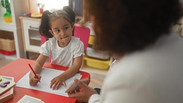 Girl drawing with pencil at red table in preschool classroom, teacher guiding hand over paper while pointing to shapes; focused learning.