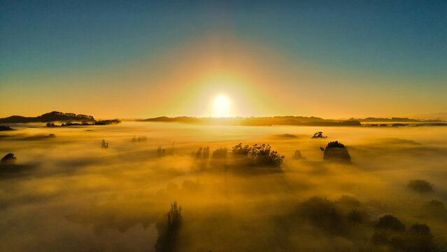 Aerial view of Lake Ratapiko at sunrise, with golden light illuminating the dense morning fog covering the landscape and surrounding trees in Ratapiko, Taranaki Region, New Zealand.