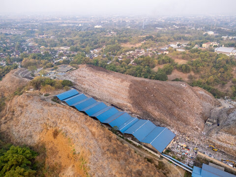 Aerial view of a waste management facility with blue industrial roofs situated in a large landfill site surrounded by hilly terrain and vegetation in Surakarta, Central Java, Indonesia.
