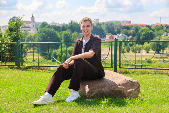 Handsome 20-year-old young man sitting on a large stone in a beautiful green park outdoors relaxing and enjoying peaceful nature scenery on sunny day.
