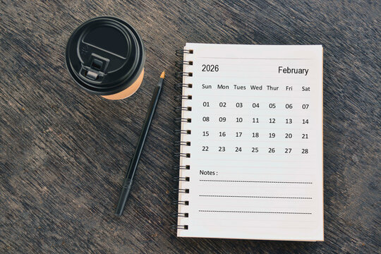 February 2026 desk calendar with disposable coffee cup and pen on wooden table. Directly above. Flat lay.
