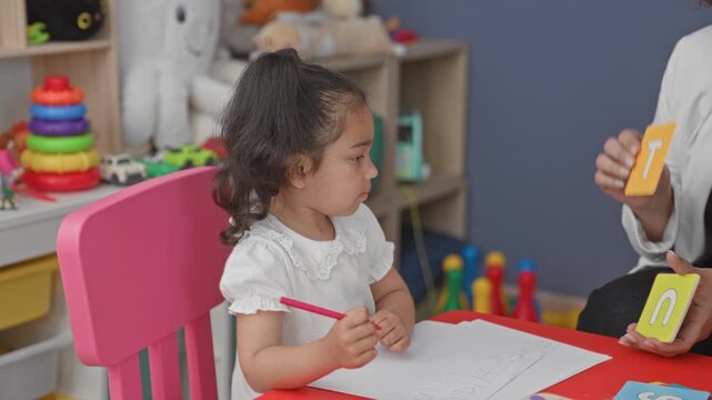 Little girl writing with pencil, hand on paper and teacher holding letter flashcards in a preschool building; concentration early learning.