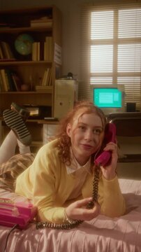 Vertical shot of Caucasian teenage girl lying on bed, blowing bubble gum and talking on retro pink telephone. Clip is ideal for nostalgia marketing campaigns and youth lifestyle commercials