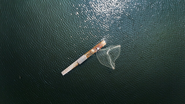 Aerial view of a fisherman casting a net from a wooden raft on the calm waters of Waduk Cirata in Cianjur, West Java, Indonesia.