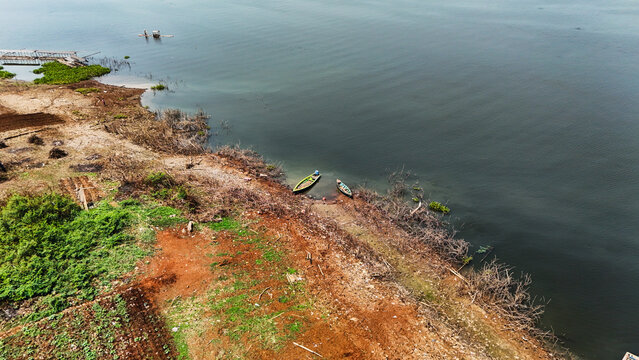 Aerial view of Jangari reservoir with small wooden boats moored along the shore, surrounded by dry vegetation and patches of green land in Cianjur, West Java, Indonesia.