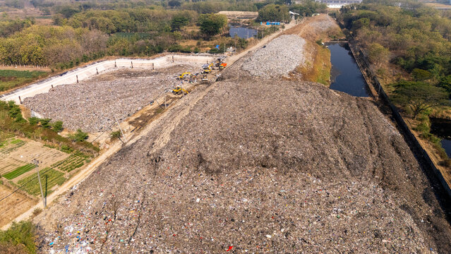Aerial view of a large landfill site with excavators managing waste piles near a water canal and agricultural fields in Klaten, Central Java, Indonesia.