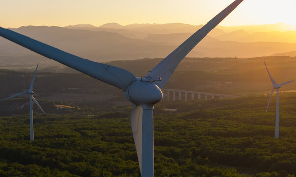 Aerial view of wind turbines illuminated by golden sunrise light, surrounded by forest and layered mountain ranges in Marsanne, Auvergne-Rhone-Alpes, France.