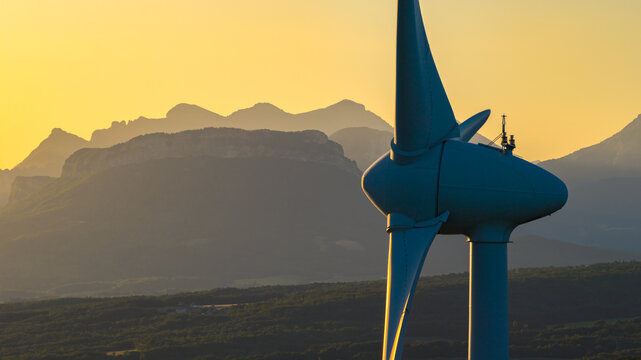 Aerial view of a wind turbine illuminated by warm sunrise light, with layered mountain silhouettes in the background, renewable energy infrastructure in Marsanne, Auvergne-Rhone-Alpes, France.