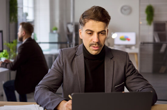 Businessman in suit in office using laptop. The manager stays serious and focused, working on tasks for corporate strategy, analysis, leadership and finance. Clear focus and productivity theme.