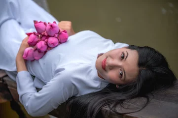 Fotobehang Lotusbloem Stunning Vietnamese woman in white traditional Ao Dai dress lying peacefully on a wooden rowboat holding a pink lotus blossom   © Jack Tamrong