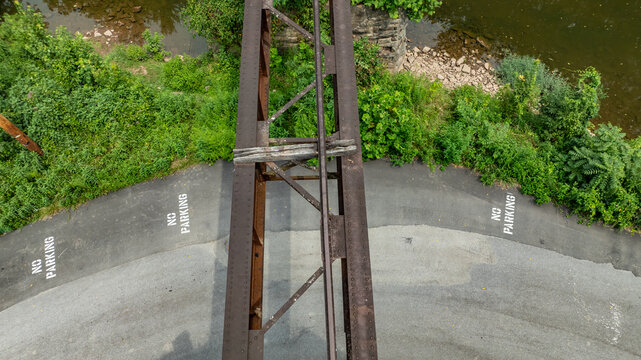 A view of an old railroad bridge looking down from above. The surrounding area has a river and green vegetation. No parking signs are visible on the road below the bridge.