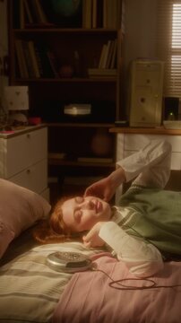 Vertical of Caucasian teenager lying on bed, pressing play on retro CD player and listening to music. Footage is suitable for nostalgia marketing and youth lifestyle campaigns