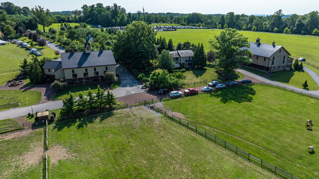 Groups of cars are parked near buildings on a farm. The land is open with green grass and trees around. It is a sunny day with clear skies and people enjoying outdoor activities.