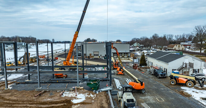 Workers use cranes and machinery to build a structure at a construction site. Snow covers the ground and nearby buildings are visible. The area shows signs of active construction.