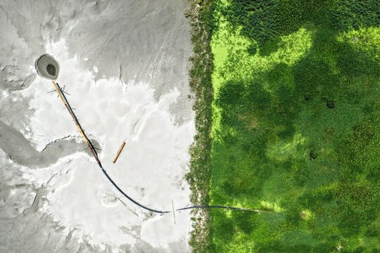 Aerial view of the Aminbazar Landfill showing the contrast between the gray industrial waste area and adjacent green wetlands with a drainage pipe in Aminbazar, Dhaka Division, Bangladesh.
