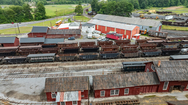 Freight cars are lined up in a train yard surrounded by several red buildings. Green fields and distant trees are visible in the background on a clear day.