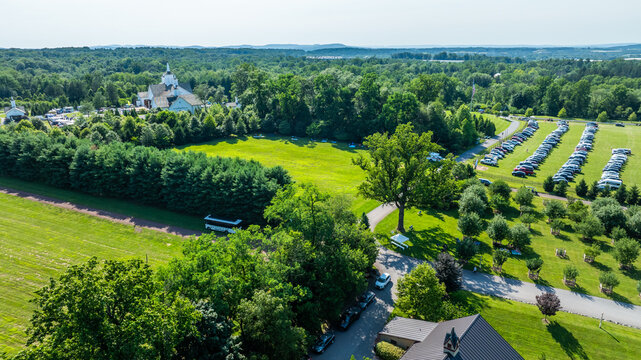 An aerial view shows a green space with many trees and a large area for parking cars. A building is visible in the background with a few hills and trees.