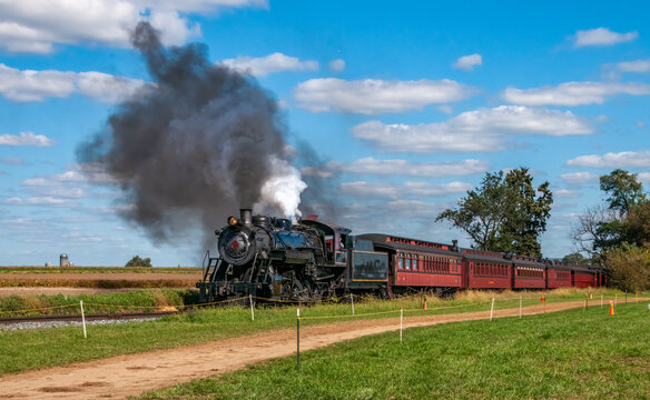 A steam train moves along the tracks surrounded by fields and trees. The train releases smoke as it journeys through the open landscape on a clear day.