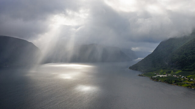Aerial view of Norddalsfjorden with dramatic sunbeams breaking through heavy clouds over the water, surrounded by steep mountains and green slopes in Valldal, More og Romsdal, Norway.