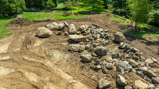 Workers are clearing an area of land where many large rocks are scattered. The scene shows dirt and grass under a clear blue sky. Trees surround the site, and it is bright outside.