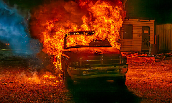 A truck is on fire in a parking lot at night. Flames and smoke are rising as the vehicle burns. Surrounding areas are dark, and the scene looks intense.