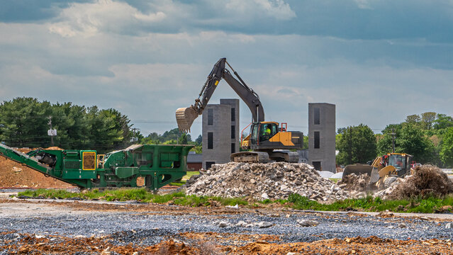 Heavy machinery operates at a construction site where workers prepare for new building development. Large equipment moves earth and materials around the area. Trees line the background.