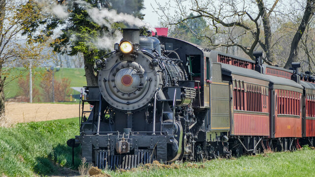 A steam train moves along the tracks surrounded by green fields. The sun shines, and trees line the way. The train releases steam as it goes by, creating a sense of movement and energy.