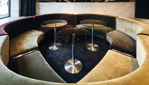 A deep square conversation pit with low curved seating, brushed chrome details, smoked-glass tables, and rich contemporary velvet upholstery in earthy jewel tones.
