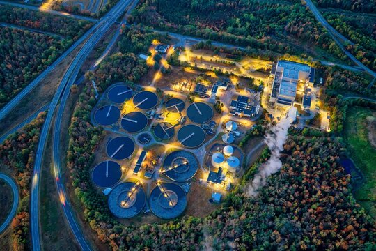 Aerial view of a wastewater treatment plant illuminated at night with circular sedimentation tanks and industrial buildings surrounded by forest near a highway in Sausheim, Grand Est, France.