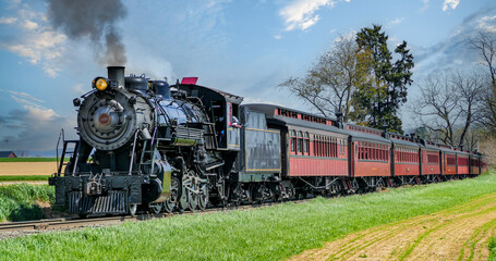 A steam train moves down a track lined with bright green grass and trees. Smoke rises from the train as it passes by a field in the background. The scene captures a moment of railway history. © Greg Kelton