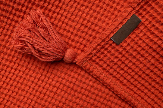 Red orange throw blanket with decorative tassels and blank fabric label appears closeup. Woven texture, soft cloth, and empty tag support product mockups, custom branding, home decor, and retail.