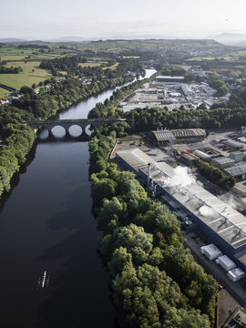 Aerial view of the River Lune in Lancaster, featuring a stone railway bridge, an industrial facility with steam, and a rowing boat on the water in Lancaster, England, United Kingdom.