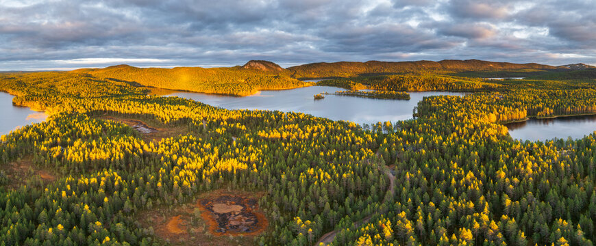 Aerial view of Hakojarvi lake surrounded by dense coniferous forests and rolling hills under a dramatic cloudy sky during golden hour in Kuusamo, Pohjois-Pohjanmaa, Finland.