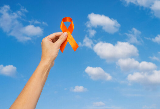 A hand holding an orange awareness ribbon isolated against a bright blue sky with soft white clouds representing support and hope