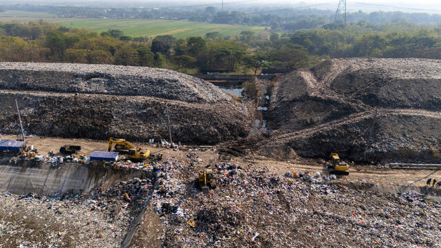 Aerial view of a large landfill site with excavators working on piles of waste surrounded by trees and fields in Klaten, Central Java, Indonesia.