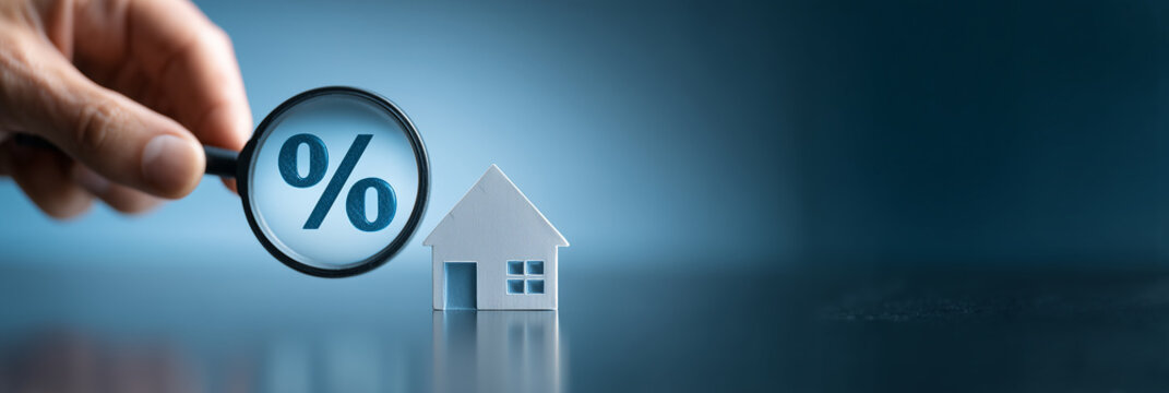 Hand holding magnifying glass over percent sign next to small house model on dark blue background. Mortgage interest rate search concept.