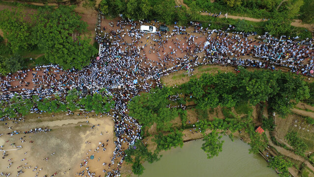 Cox's Bazar, Bangladesh - 25 August 2026: Aerial view of a large crowd gathered at the Rohingya refugee camp, with people walking along dirt paths near a pond and green trees.
